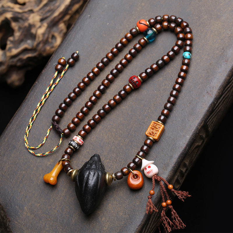 Full view of Tibetan Nala Tsa Tsa mala necklace laid on dark wood surface showing aged brown beads, antique glass, skull ornaments and black pendant with tassels