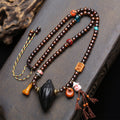 Full view of Tibetan Nala Tsa Tsa mala necklace laid on dark wood surface showing aged brown beads, antique glass, skull ornaments and black pendant with tassels