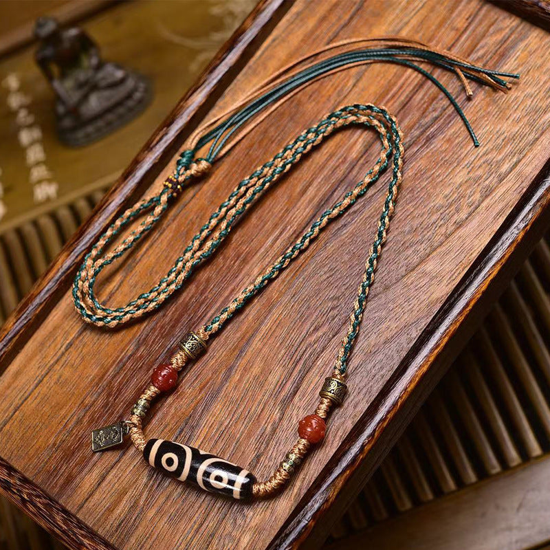 Full view of hand-woven two-tone green and tan cord necklace with three-eye dzi bead pendant laid flat on wooden surface with tassel ends
