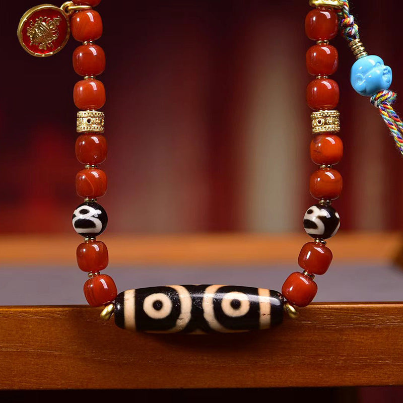 Three-eye dzi bead centered on red agate necklace with decorative brass charms and colorful braided tassel