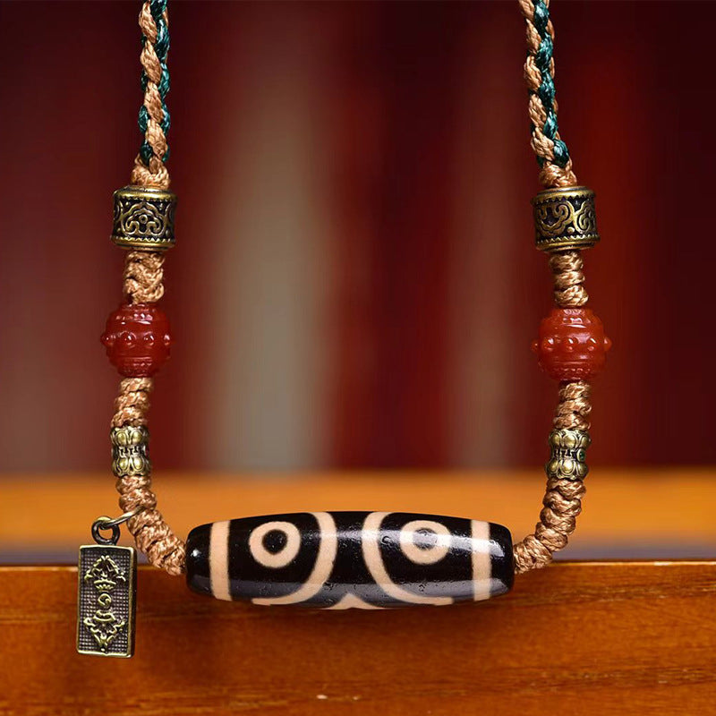 Front view of three-eye dzi bead centered on two-tone braided cord with red agate beads and antique bronze metal ornaments and vintage tag charm