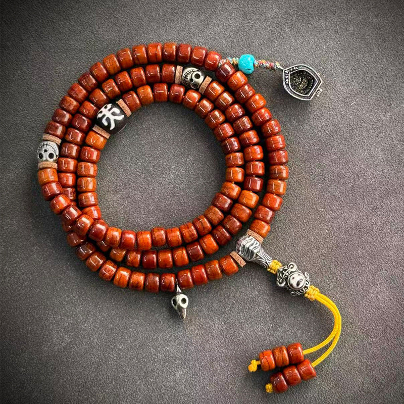 Yak bone mala coiled on grey surface with dzi bead, red agate, crystal drop and colorful tassel